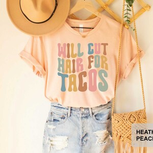 May include: A light pink t-shirt with the text "Will Cut Hair For Tacos" in colorful retro font. The shirt is being modeled on a person wearing denim shorts and a straw hat.