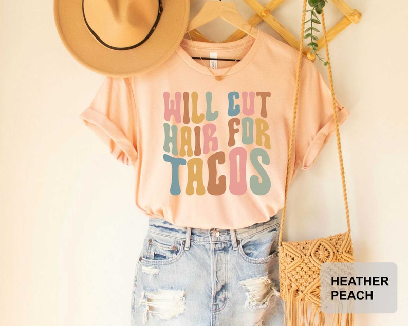 May include: A light pink t-shirt with the text "Will Cut Hair For Tacos" in colorful retro font. The shirt is being modeled on a person wearing denim shorts and a straw hat.
