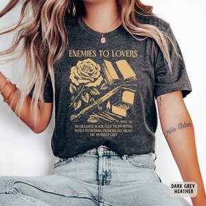 May include: Dark grey heather t-shirt with a graphic design of a rose, a book, a dagger, and matches. The text "Enemies to Lovers" and "Issue 02" are printed on the shirt. The text "An exclusive book club from bitter rivals to burning passions all about the morally grey" is also printed on the shirt.