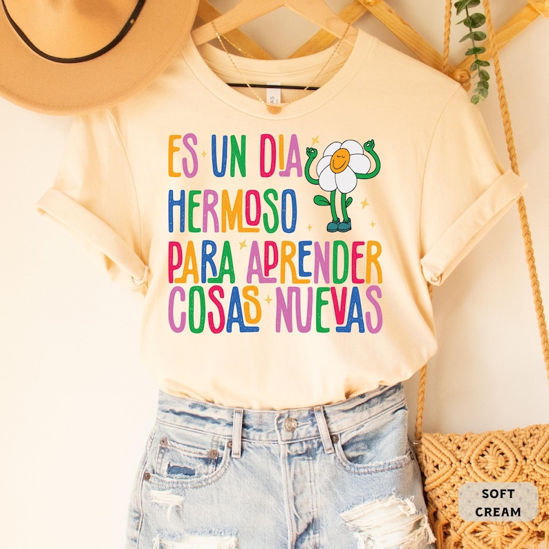 May include: A cream colored t-shirt with a colorful graphic that says "Es un dia hermoso para aprender cosas nuevas" and a cartoon flower with arms and legs.