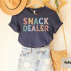 Puede incluir: Camiseta azul marino con el texto "SNACK DEALER" en letras de colores. La camiseta se muestra en una modelo que lleva pantalones cortos de mezclilla y un bolso de paja.