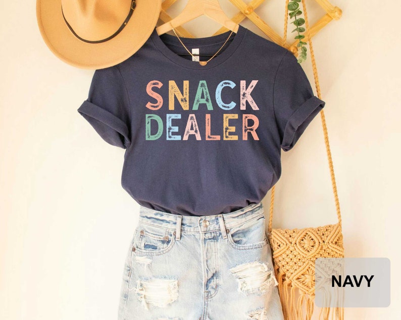Puede incluir: Camiseta azul marino con el texto "SNACK DEALER" en letras de colores. La camiseta se muestra en una modelo que lleva pantalones cortos de mezclilla y un bolso de paja.