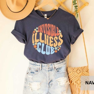 K&ouml;nnte beinhalten: Ein marineblaues T-Shirt mit einer bunten Grafik, auf der in einer Retro-Schrift "Invisible Illness Club" steht. Die Grafik ist von kleinen Blumenmustern umgeben. Das Shirt wird von einer Person getragen, die eine Jeans-Shorts tr&auml;gt.