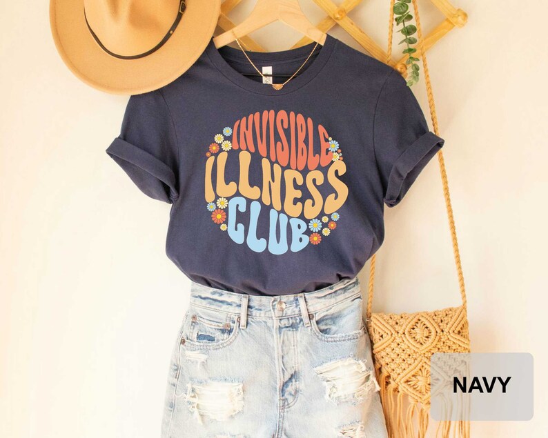 K&ouml;nnte beinhalten: Ein marineblaues T-Shirt mit einer bunten Grafik, auf der in einer Retro-Schrift "Invisible Illness Club" steht. Die Grafik ist von kleinen Blumenmustern umgeben. Das Shirt wird von einer Person getragen, die eine Jeans-Shorts tr&auml;gt.