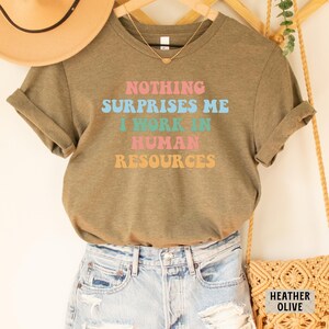 May include: A green t-shirt with the text "NOTHING SURPRISES ME I WORK IN HUMAN RESOURCES" in colorful retro font.