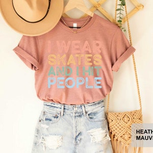 May include: A heather mauve t-shirt with the text "I wear skates and I hit people" in rainbow colours. The shirt is displayed on a model wearing denim shorts and a straw hat.