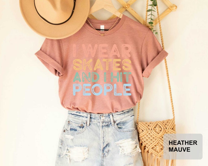May include: A heather mauve t-shirt with the text "I wear skates and I hit people" in rainbow colours. The shirt is displayed on a model wearing denim shorts and a straw hat.