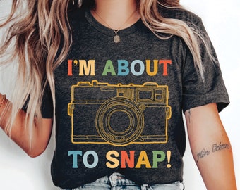 May include: A dark gray t-shirt with the text "I'M ABOUT TO SNAP!" in rainbow colors. A vintage camera illustration is above the text.