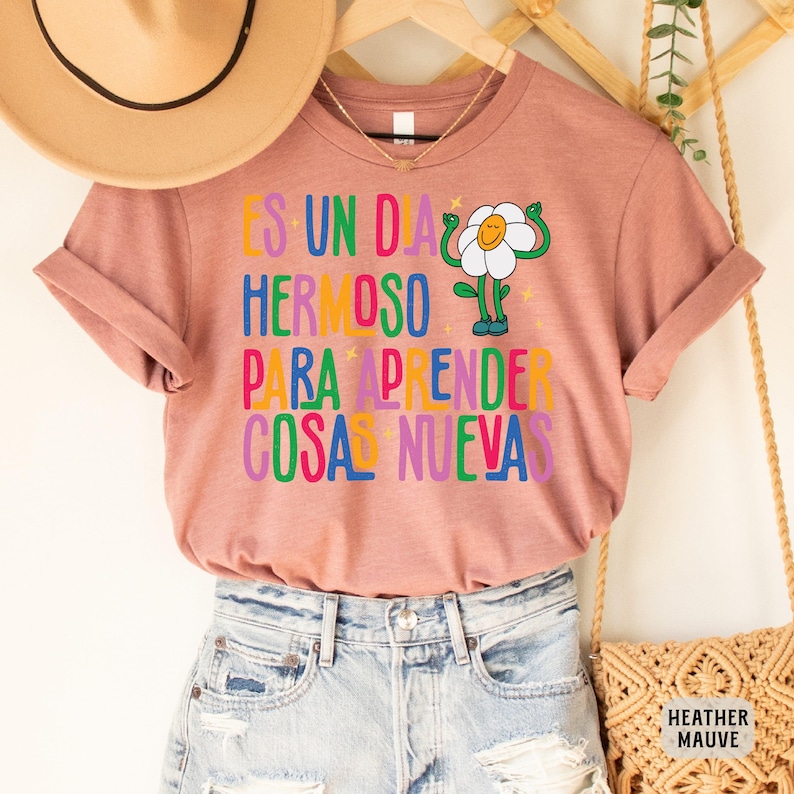 May include: A heather mauve t-shirt with the text "Es un dia hermoso para aprender cosas nuevas" in colorful letters. A cartoon flower with a smiling face and arms raised in the air is next to the text.