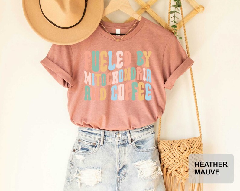 Op de afbeelding: Een heide-paarse t-shirt met de tekst "Fueled by Mitochondria and Coffee" in een retro-lettertype met kleurrijke letters. Het shirt wordt getoond op een mannequin met een strohoed en een spijkerrok.