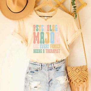 May include: A light beige t-shirt with the text "Psychology Major Cuz Every Family Needs A Therapist" in colorful, retro-style lettering.