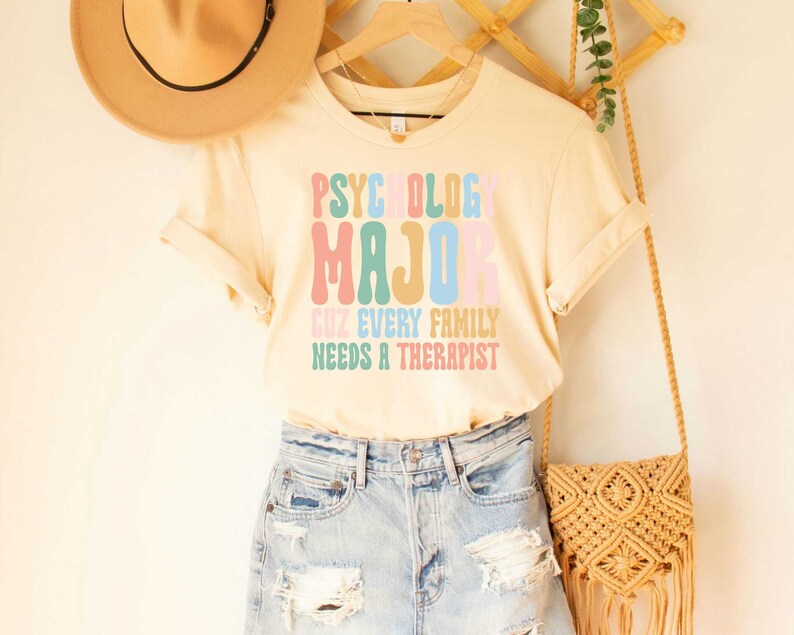 May include: A light beige t-shirt with the text "Psychology Major Cuz Every Family Needs A Therapist" in colorful, retro-style lettering.