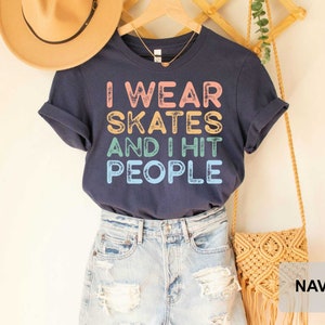 May include: A navy blue t-shirt with the text "I wear skates and I hit people" in rainbow colours. The shirt is shown on a model wearing denim shorts.