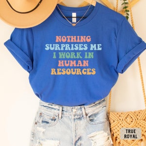 May include: A royal blue t-shirt with the text "Nothing surprises me I work in human resources" in a retro font with colorful letters.