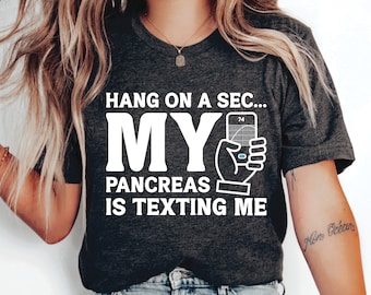 May include: Dark gray t-shirt with the text "Hang on a sec... My Pancreas is Texting Me" in white. The graphic includes a hand holding a blood glucose meter. The shirt is a crew neck.