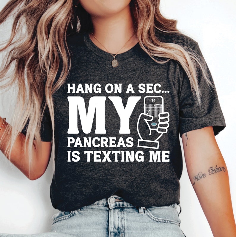 May include: Dark gray t-shirt with the text "Hang on a sec... My Pancreas is Texting Me" in white. The graphic includes a hand holding a blood glucose meter. The shirt is a crew neck.