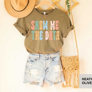 May include: A heather olive green t-shirt with the text "Show Me The Data" in colorful retro font. The shirt is hanging on a wooden hanger with a straw hat and a macrame bag.