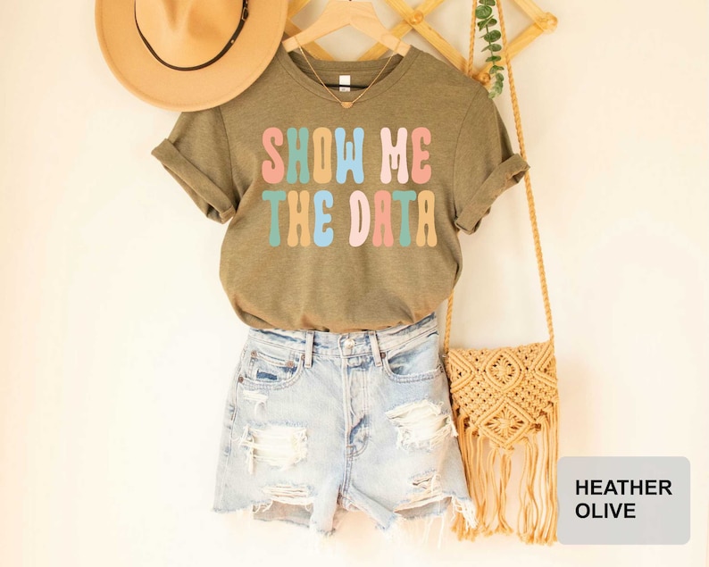 May include: A heather olive green t-shirt with the text "Show Me The Data" in colorful retro font. The shirt is hanging on a wooden hanger with a straw hat and a macrame bag.