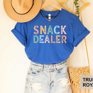 Puede incluir: Una camiseta azul real con el texto "Snack Dealer" en colores arco&iacute;ris. La camiseta se lleva con pantalones cortos de mezclilla y un bolso de paja.