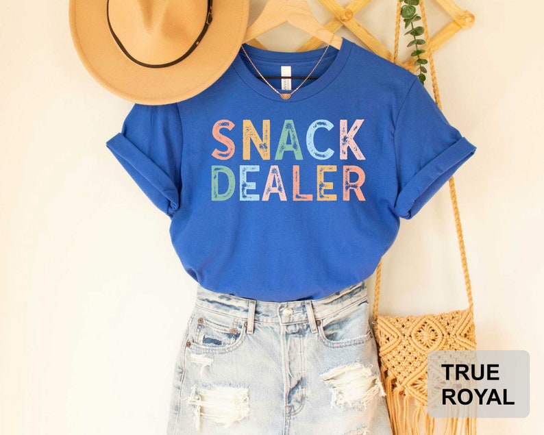 Puede incluir: Una camiseta azul real con el texto "Snack Dealer" en colores arco&iacute;ris. La camiseta se lleva con pantalones cortos de mezclilla y un bolso de paja.