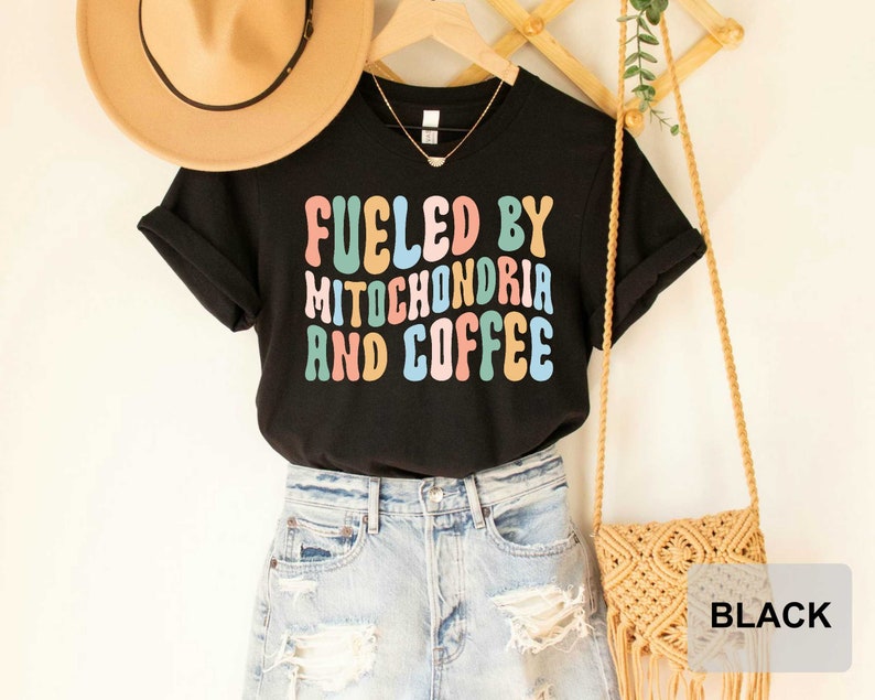 Op de afbeelding: Zwart T-shirt met de tekst "Fueled by Mitochondria and Coffee" in kleurrijke, retro-stijl letters.