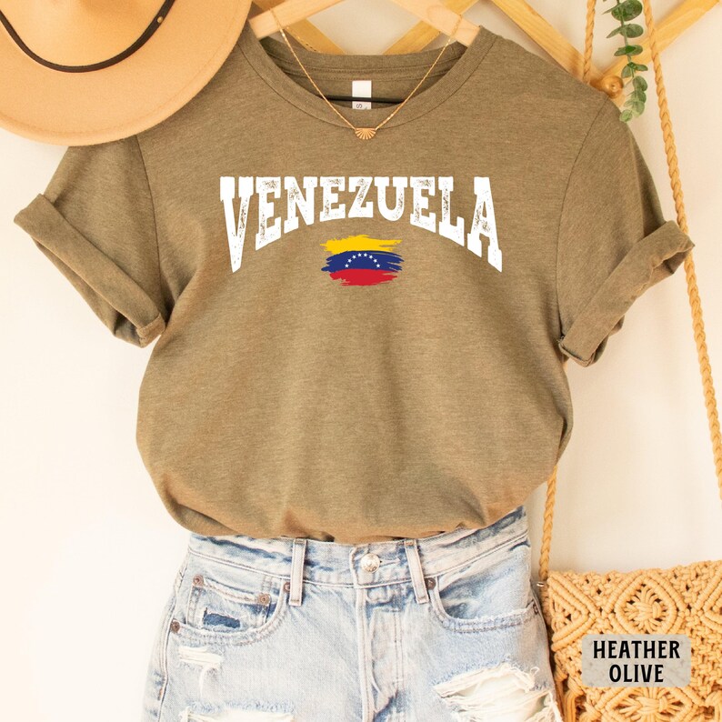 May include: A heather olive t-shirt with the word "VENEZUELA" in white letters with a distressed look. The shirt also features a stylized image of the Venezuelan flag.