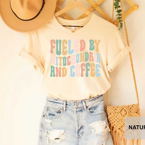 Op de afbeelding: Een licht beige T-shirt met de tekst "Fueled by Mitochondria and Coffee" in kleurrijke, retro-stijl letters.