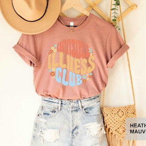 K&ouml;nnte beinhalten: Heather-mauvefarbenes T-Shirt mit dem Text "Invisible Illness Club" in einer Retro-Schriftart mit bunten Blumen, die den Text umgeben. Das Shirt wird von einer Person mit Jeans-Shorts und einem Strohhut getragen.