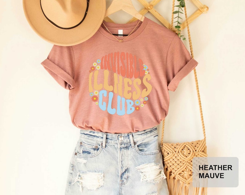 K&ouml;nnte beinhalten: Heather-mauvefarbenes T-Shirt mit dem Text "Invisible Illness Club" in einer Retro-Schriftart mit bunten Blumen, die den Text umgeben. Das Shirt wird von einer Person mit Jeans-Shorts und einem Strohhut getragen.