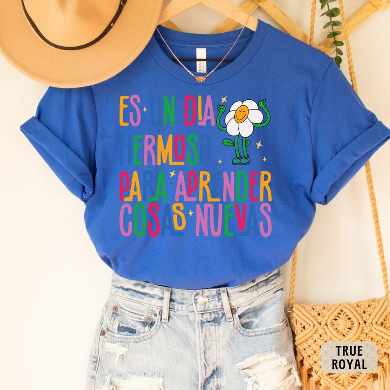 May include: A royal blue t-shirt with a colorful graphic that says "Es un dia hermoso para aprender cosas nuevas" and a cartoon flower with arms and legs.