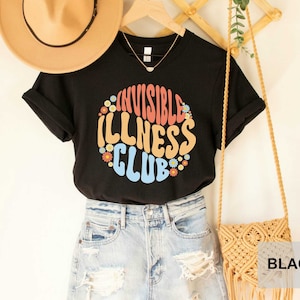 K&ouml;nnte beinhalten: Schwarzes T-Shirt mit dem Text "Invisible Illness Club" in einer Retro-Schriftart mit bunten Blumen, die den Text umgeben.