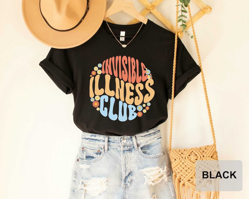 K&ouml;nnte beinhalten: Schwarzes T-Shirt mit dem Text "Invisible Illness Club" in einer Retro-Schriftart mit bunten Blumen, die den Text umgeben.