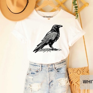 May include: White t-shirt with a black and white illustration of a raven on the front. The shirt is being modeled on a person wearing blue jeans and a brown hat.