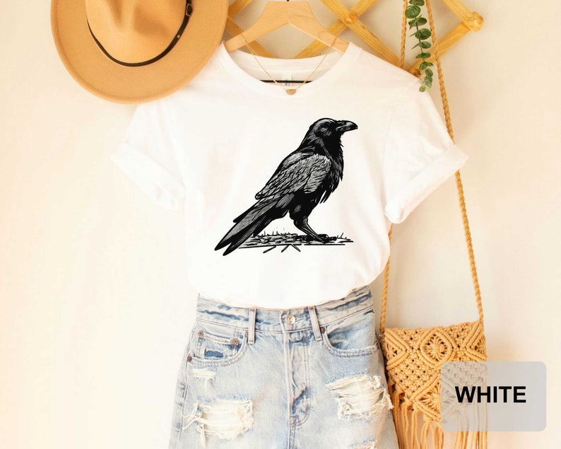 May include: White t-shirt with a black and white illustration of a raven on the front. The shirt is being modeled on a person wearing blue jeans and a brown hat.