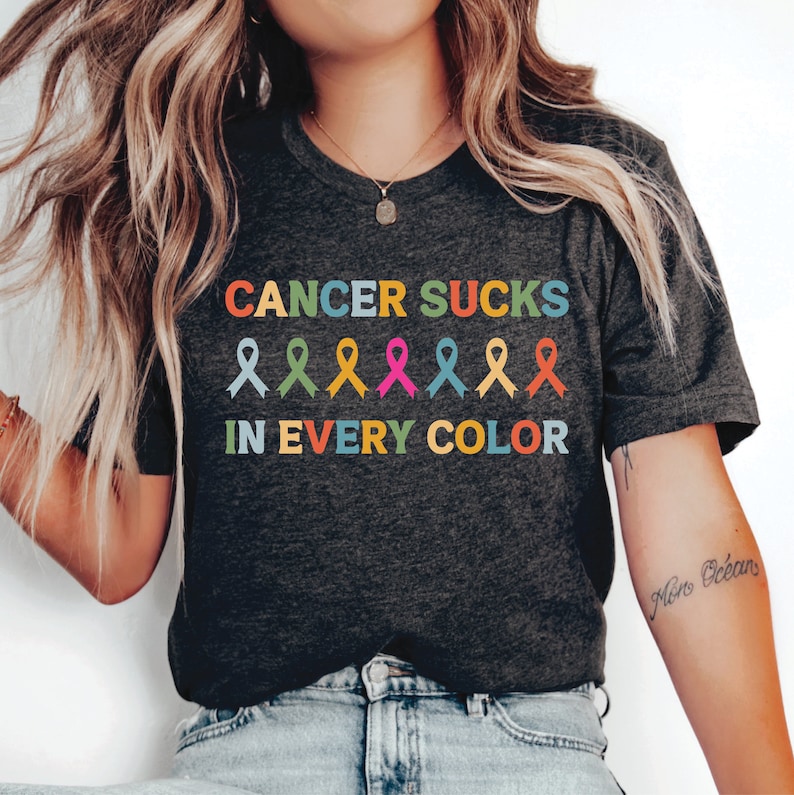 May include: A dark grey t-shirt with the text "Cancer Sucks in Every Color" in rainbow colors. The text is surrounded by a row of colorful ribbon symbols.