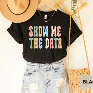 May include: Black t-shirt with the text "SHOW ME THE DATA" in colorful, retro-style lettering. The shirt is worn with a pair of light blue denim shorts and a brown woven bag.