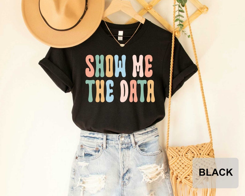 May include: Black t-shirt with the text "SHOW ME THE DATA" in colorful, retro-style lettering. The shirt is worn with a pair of light blue denim shorts and a brown woven bag.