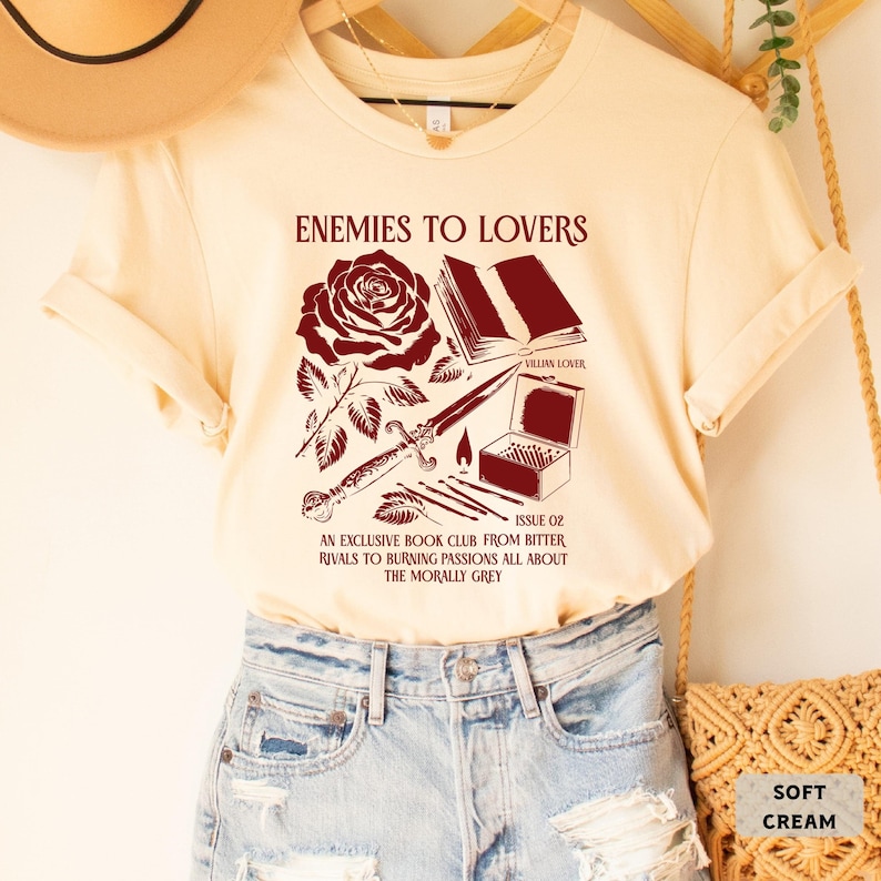 May include: A cream colored t-shirt with a maroon graphic design featuring a rose, a book, a dagger, matches, and a box. The text on the shirt reads "Enemies to Lovers", "Villain Lover", "Issue 02", "An Exclusive Book Club From Bitter Rivals to Burning Passions All About The Morally Grey", and "Soft Cream".