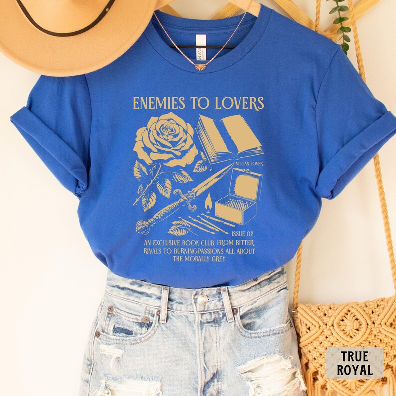 May include: A royal blue t-shirt with a gold graphic design featuring a rose, a book, a dagger, and matches. The text on the shirt reads "Enemies to Lovers" and "An exclusive book club from bitter rivals to burning passions all about the morally grey".