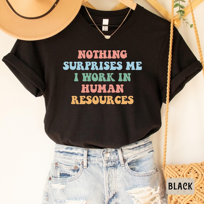 May include: Black t-shirt with the text "Nothing surprises me I work in human resources" in colorful retro font.
