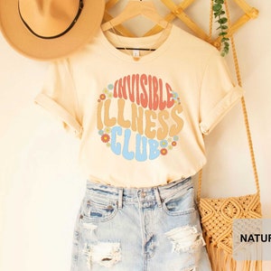 K&ouml;nnte beinhalten: Ein beiges T-Shirt mit einer Retro-Grafik, auf der "Invisible Illness Club" in Regenbogenfarben mit Blumen um den Text steht. Das Shirt wird von einer Person mit Jeans-Shorts und einem Strohhut getragen.