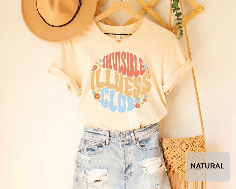 K&ouml;nnte beinhalten: Ein beiges T-Shirt mit einer Retro-Grafik, auf der "Invisible Illness Club" in Regenbogenfarben mit Blumen um den Text steht. Das Shirt wird von einer Person mit Jeans-Shorts und einem Strohhut getragen.