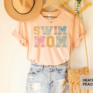 Pu&ograve; includere: Una maglietta color pesca con il testo "Swim Mom" in un font in stile vintage con accenti blu, rosa e gialli. La maglietta &egrave; indossata con un cappello di paglia e una borsa all'uncinetto.