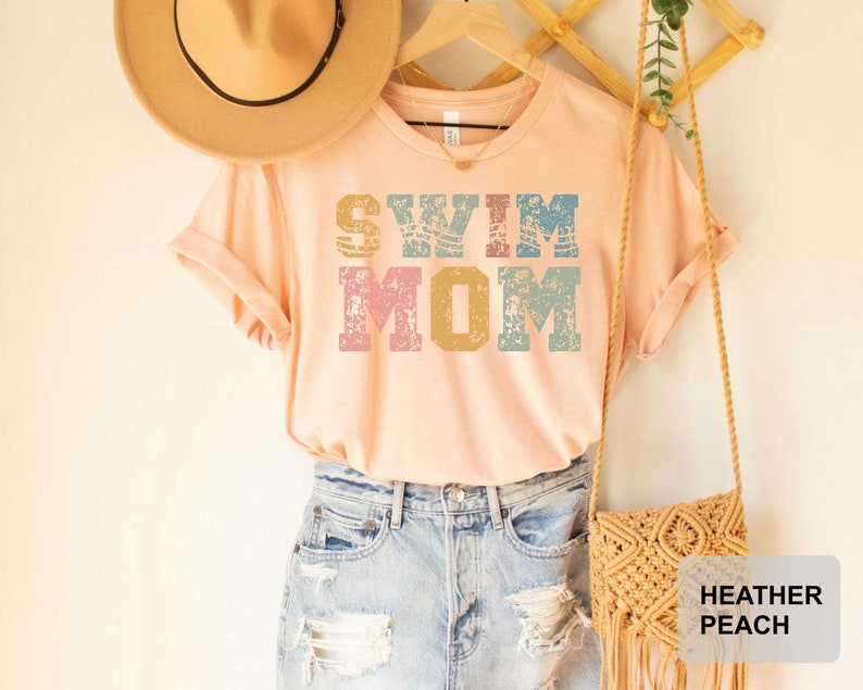 Pu&ograve; includere: Una maglietta color pesca con il testo "Swim Mom" in un font in stile vintage con accenti blu, rosa e gialli. La maglietta &egrave; indossata con un cappello di paglia e una borsa all'uncinetto.