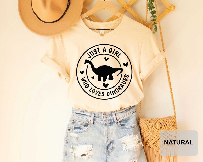 Puede incluir: Una camiseta beige con un gr&aacute;fico negro que dice "Just a girl who loves dinosaurs" con una silueta de dinosaurio y corazones. La camiseta est&aacute; en una percha y tiene un sombrero marr&oacute;n y una bolsa beige en el fondo.