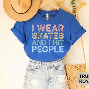 May include: Blue t-shirt with the text "I wear skates and I hit people" in rainbow colours. The shirt is being modelled on a person wearing blue jeans and a brown hat.