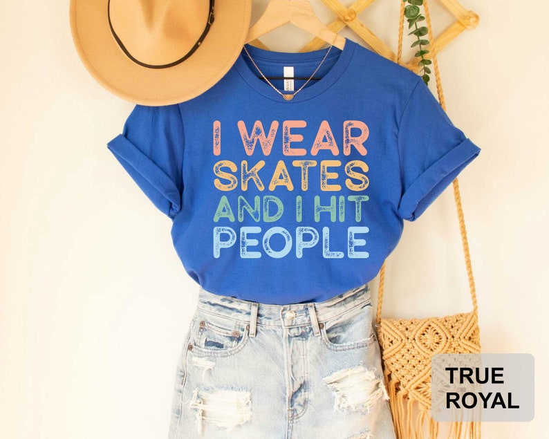 May include: Blue t-shirt with the text "I wear skates and I hit people" in rainbow colours. The shirt is being modelled on a person wearing blue jeans and a brown hat.