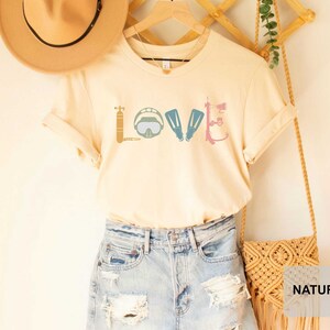 Peut inclure: Un t-shirt beige clair avec un motif graphique d'&eacute;quipement de plong&eacute;e sous-marine qui &eacute;pelle le mot "LOVE". Le t-shirt est suspendu &agrave; un cintre en bois et est associ&eacute; &agrave; une jupe en jean bleu clair et un sac tiss&eacute; naturel.