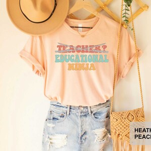 May include: A peach colored t-shirt with the text "Teacher? Educational Ninja" in a retro font. The text is in shades of blue, pink, orange, and yellow. The shirt is hanging on a wooden hanger with a straw hat and a woven bag.