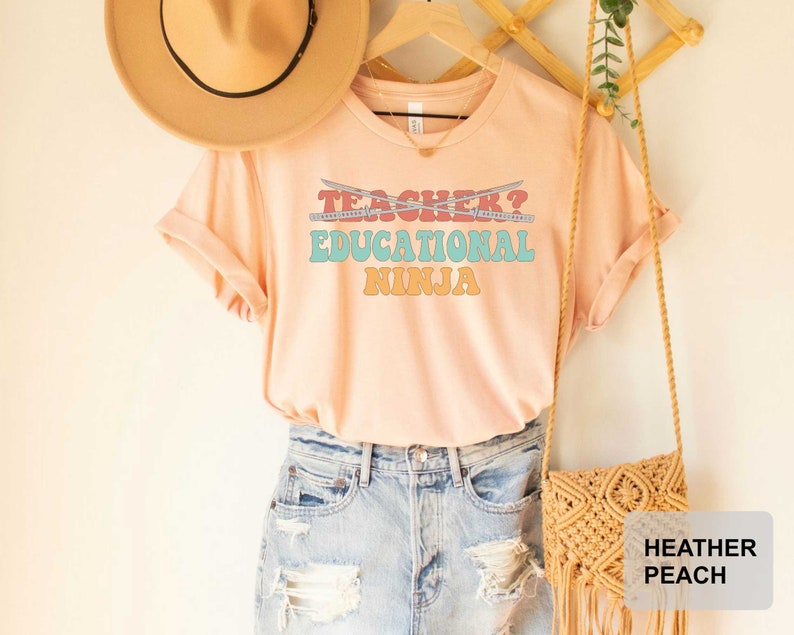 May include: A peach colored t-shirt with the text "Teacher? Educational Ninja" in a retro font. The text is in shades of blue, pink, orange, and yellow. The shirt is hanging on a wooden hanger with a straw hat and a woven bag.